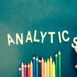 Colored pencils and letters spelling 'ANALYTICS' on a textured background with a hand.